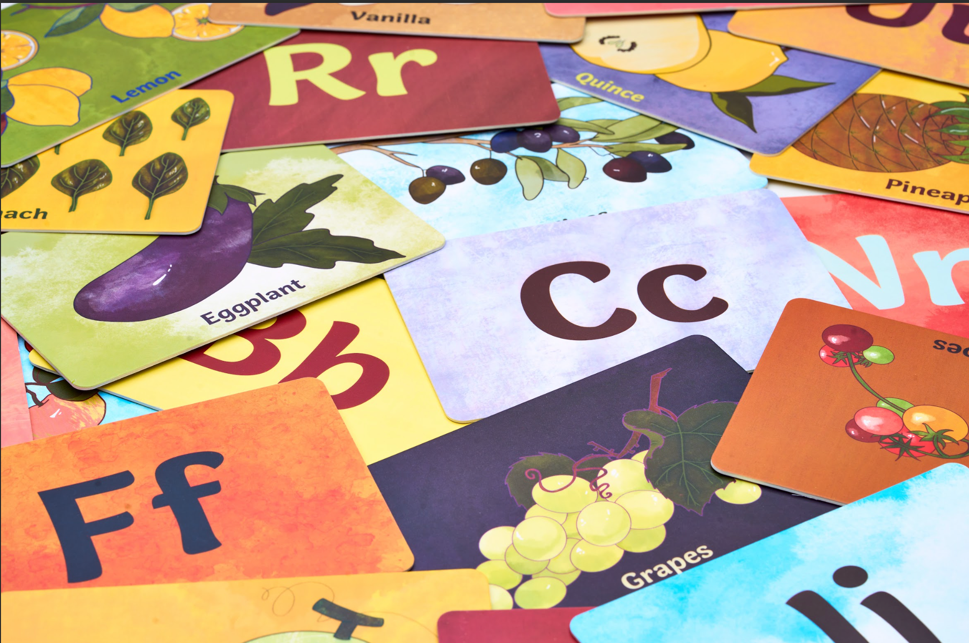 A scattered assortment of colorful alphabet flashcards featuring letters and corresponding fruits or vegetables, such as C for Grapes and E for Eggplant.