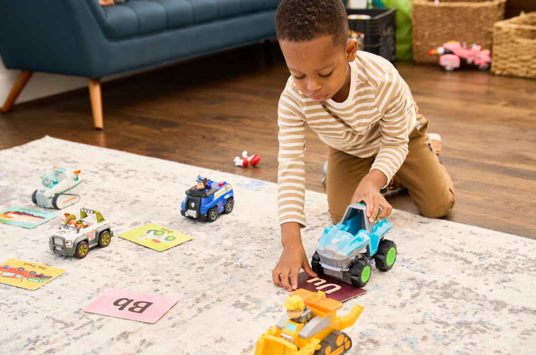 A young child plays with toy cars on a rug with scattered alphabet cards and small toys in a living room environment.
