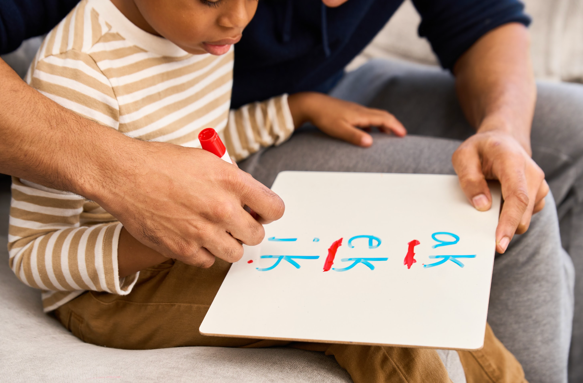 An adult helps a child write on a dry-erase board using a red marker. The child is wearing a striped shirt and light brown pants.