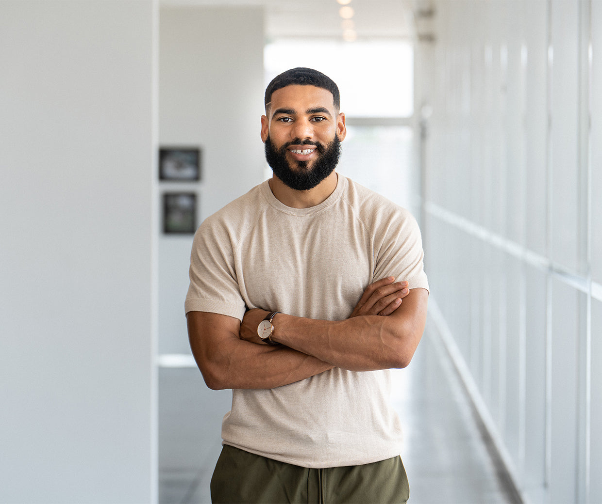 A man with a beard and short hair stands in a bright hallway with his arms crossed, wearing a beige t-shirt and green pants, smiling at the camera.