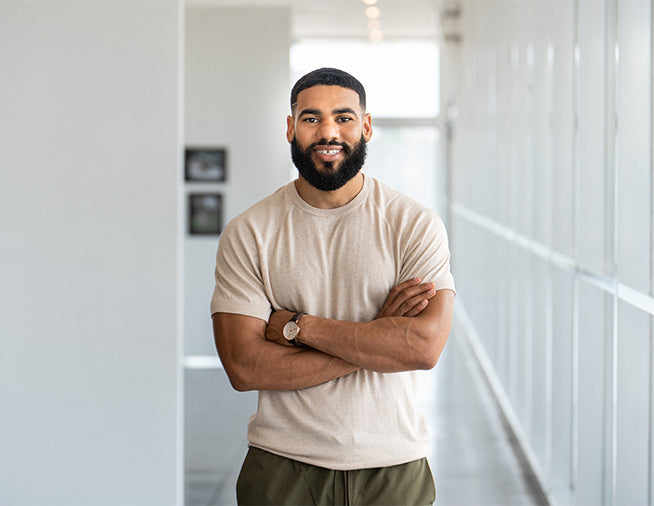 A man with a beard stands with arms crossed in a hallway, wearing a light-colored t-shirt and dark pants.