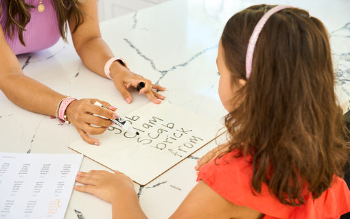 An adult assists a young girl with writing practice on a whiteboard at a marble counter. The whiteboard displays several words. A worksheet with a list of words is at the corner of the counter.