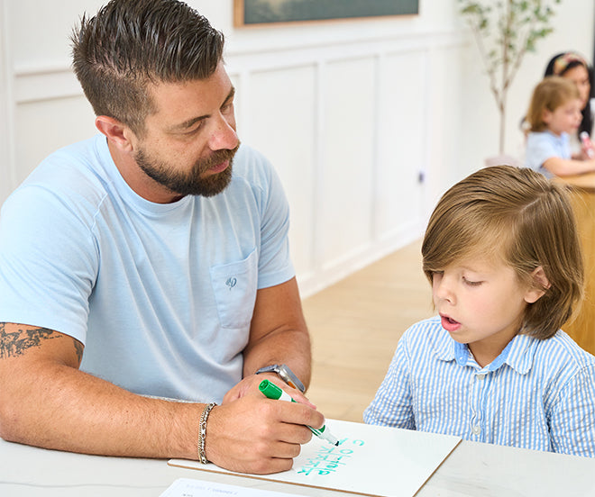 A man with a short beard and tattoos helps a young boy write on a sheet of paper with a green marker in a classroom setting. Other children and a woman are seen in the background.