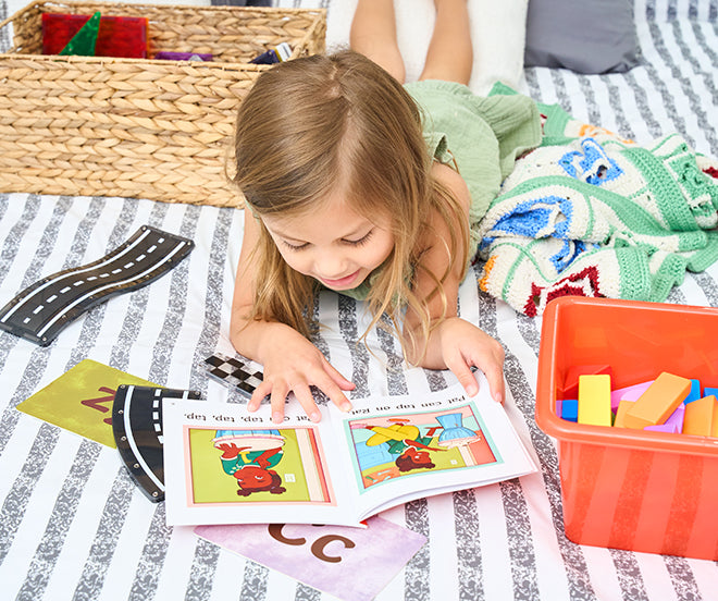 A young child lies on a striped blanket, reading a book. Surrounding the child are a woven basket, a colorful blanket, foam letters, and a road playmat.