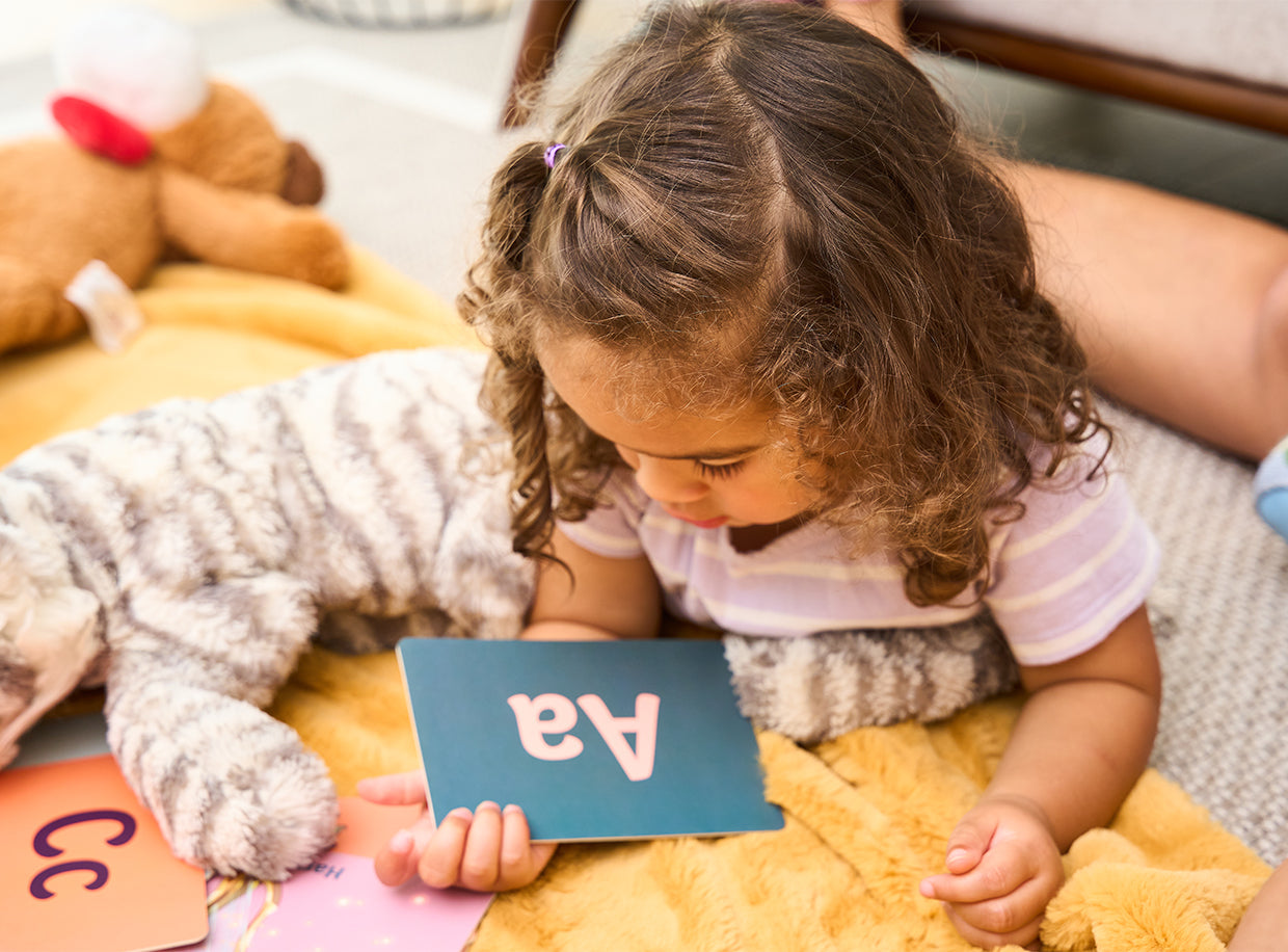 A young child lies on a yellow blanket, holding a card with the letters Aa. A stuffed animal and more letter cards are nearby.