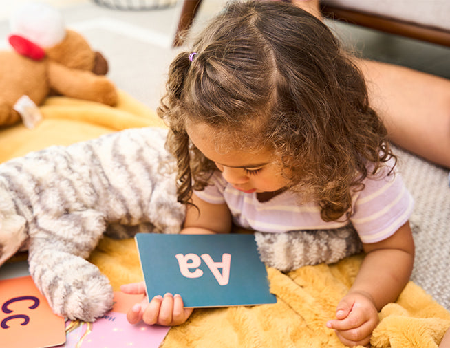 A young child with curly hair lies on a blanket, holding a flashcard with Aa printed on it, surrounded by toys and stuffed animals.