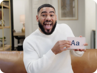 A man with a beard and white sweater holds a card displaying Aa while smiling with an open mouth in a home setting.
