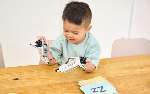 A young child sits at a wooden table, holding two toy space shuttles. Colorful flashcards are laid out on the table in front of the child.
