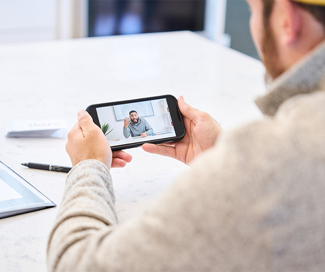 A person holds a smartphone displaying a video call with another person. A pen and an open binder are visible on the table in the foreground.