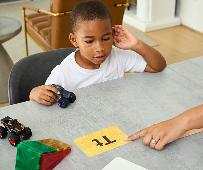 A child sits at a table holding a toy truck, looking at a card with the tt text on it while an adult points at the card.