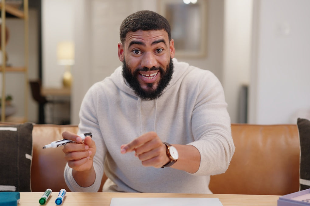 A man with a beard, wearing a light-colored sweater, is smiling and pointing while holding a whiteboard marker, sitting at a table with markers and papers.