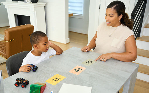 A woman and a child sit at a table. The woman points to a paper on the table featuring letters, while the child holds toy cars. A fireplace and a chair are in the background.