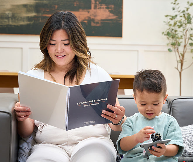 An adult and a child sit on a couch. The adult is reading a book titled Learning Sounds, while the child plays with a toy airplane.