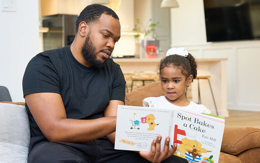 A man and a child sit on a couch reading the book Spot Bakes a Cake together. The man points to a page in the book, while the girl attentively looks on.