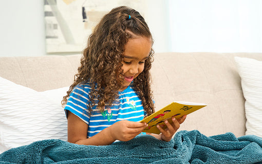 A young girl with curly hair, wearing a blue striped shirt, smiles as she reads a book while sitting on a beige couch under a green blanket.