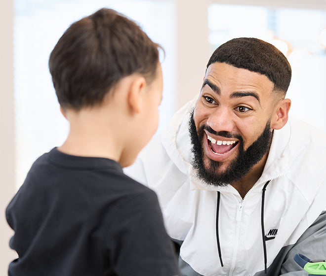 An adult with a beard and mustache smiles widely at a child who is facing away. Both are indoors with soft natural light coming in from the background.