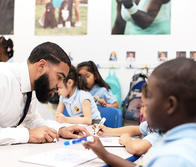 A teacher engages with young students who are drawing with colored pencils at a classroom table.