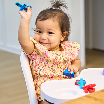 A toddler in a floral dress sits in a high chair, holding a blue toy block up in the air with a smile. More colorful blocks are placed on the high chair's tray.