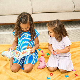 Two young children sitting on a yellow blanket; one is reading a book, while the other looks on. Colorful alphabet letters and a pink toy are scattered around them on the blanket.