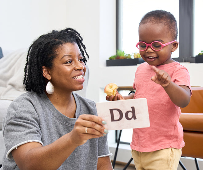 An adult shows a flashcard with the letter D to a child wearing glasses who is pointing and holding a snack.