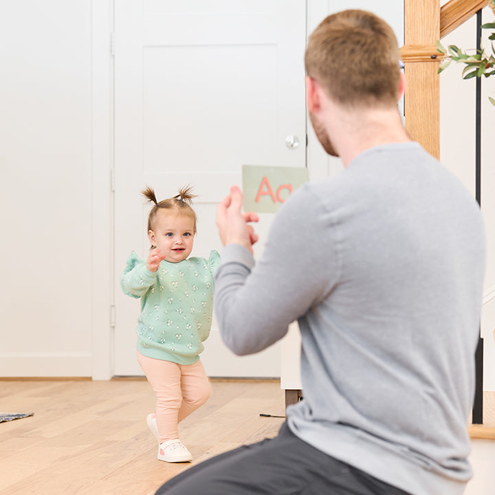 A toddler with pigtails takes steps towards an adult holding a card with A on it in a bright room with wooden flooring and a staircase in the background.