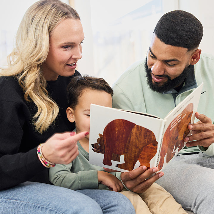 Three people, two adults and one child, sit together reading a picture book featuring a bear. The child is seated between the adults, who are looking at the book attentively.