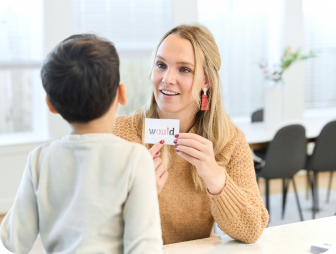 A woman holds up a flashcard with the word would while speaking to a child in a light-filled room.