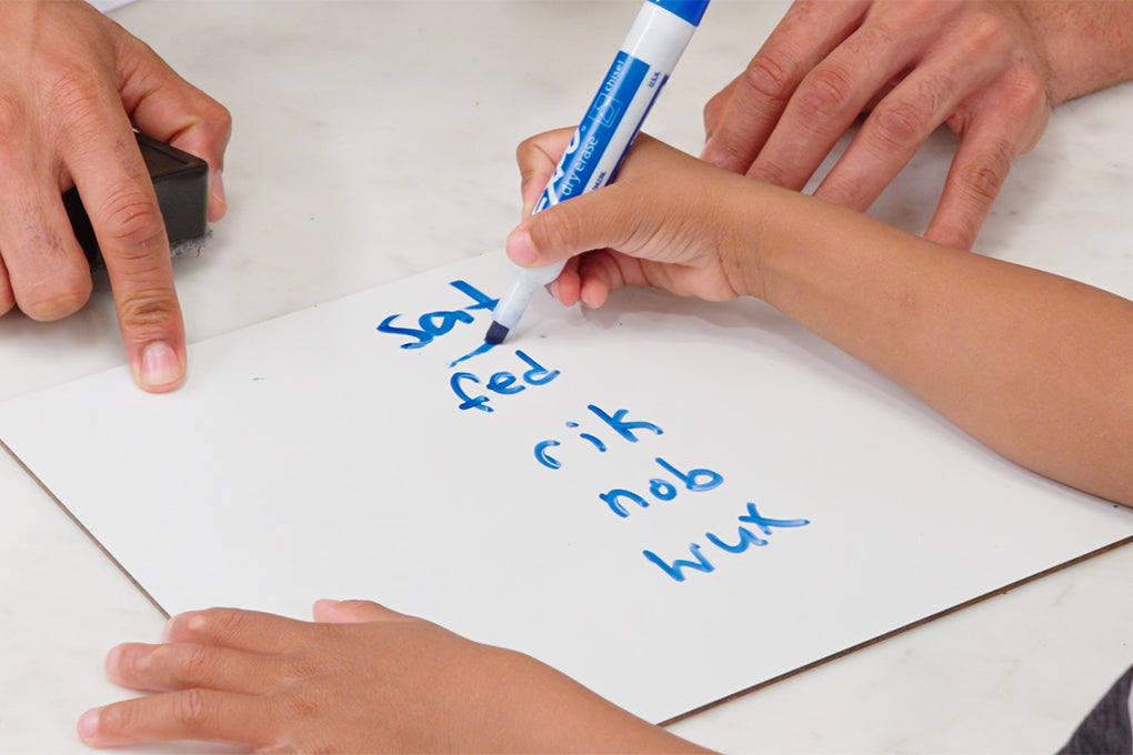A child writes a list of words in blue marker on a white board, while an adult points to the board.