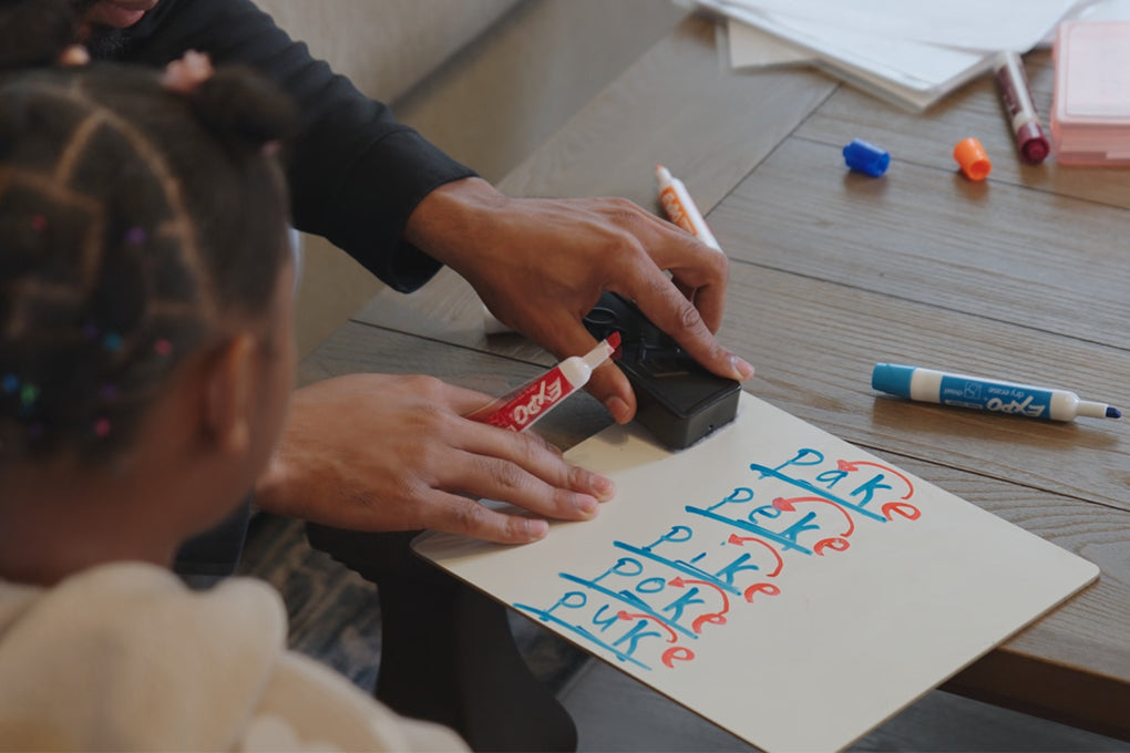 An adult helps a child write the word poke multiple times on a piece of paper using red and blue markers on a wooden table. Various markers and papers are scattered on the table.