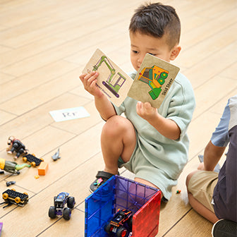 A child in a light green outfit sits on a wooden floor, holding two illustrated cards of construction vehicles. Various toy cars and construction blocks are scattered around. Another child is partially visible.