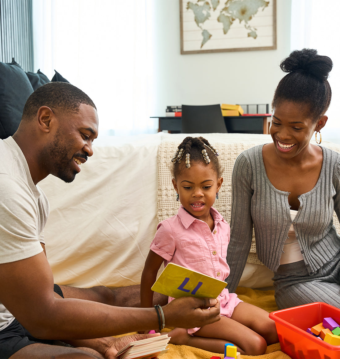 A man, woman, and young girl are sitting on the floor, engaging in an educational activity with blocks and a book in a home setting.