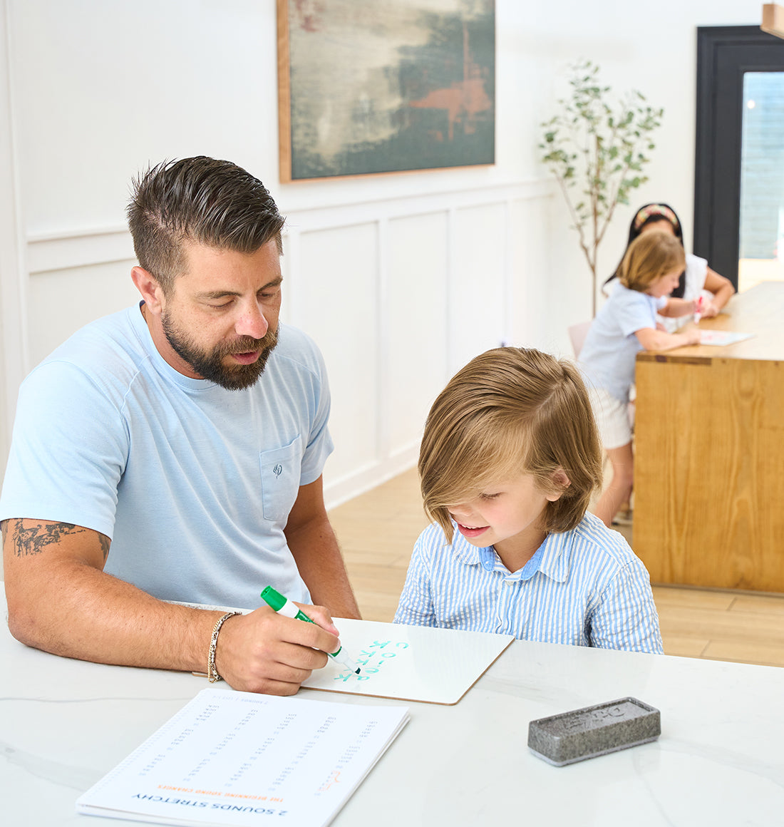An adult with a beard and tattoo teaches a child to write on a small whiteboard. In the background, another adult assists two children at a table.