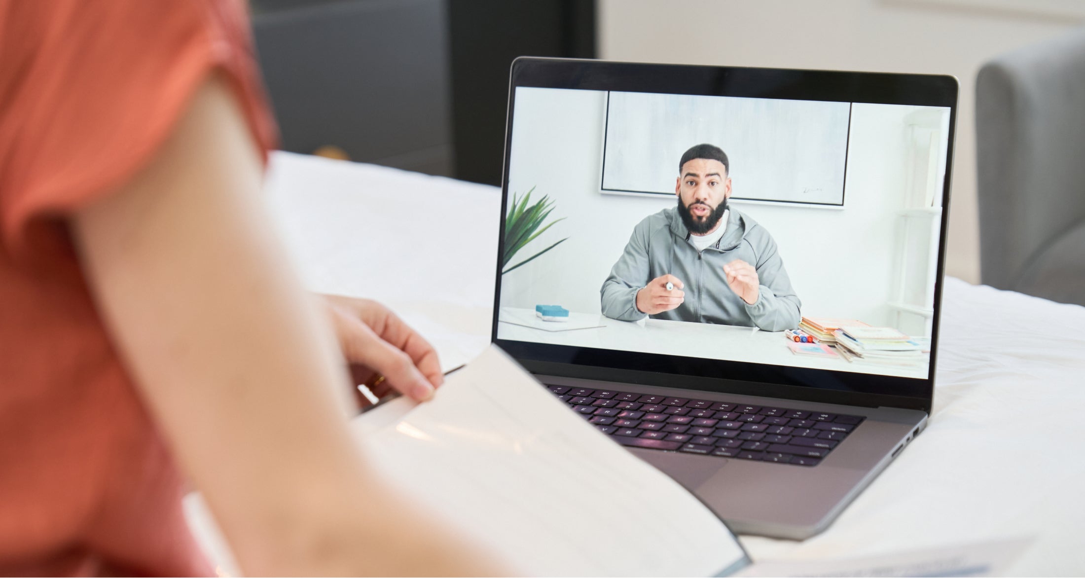 Person watching a man speaking in an online video conference on a laptop, with open documents in hand.